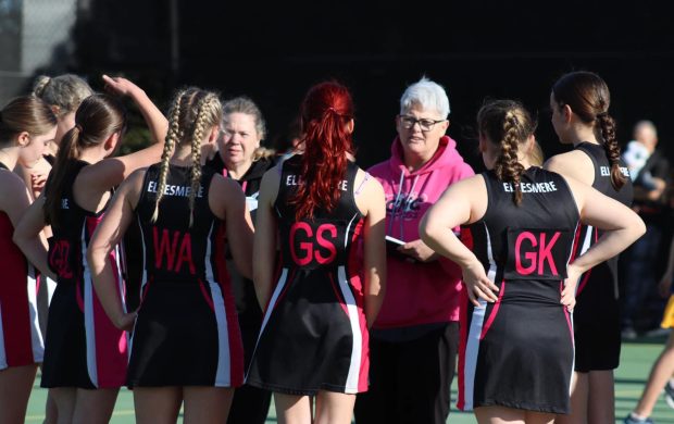 The image shows a group of female netball players in a team huddle, engaging in a discussion with their coach. The coach, wearing a bright pink hoodie, is giving instructions or feedback to the attentive players, who are dressed in matching black and pink uniforms labeled with positions like 