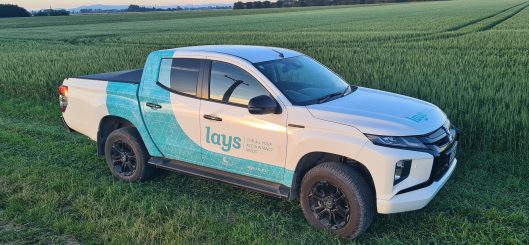 The image features a white pickup truck with blue and turquoise branding for Lay's on its side, parked in a green wheat field during what appears to be early evening. The truck, likely used for promotional or delivery purposes, is shown in a natural, rural setting, emphasizing a connection to farming or agricultural activities.