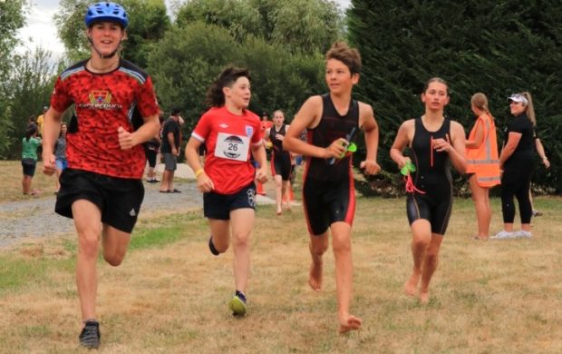 Four young athletes taking part in a Selwyn Sports triathlon. All are running. Two wearing wet suits. One with cycle helmet/